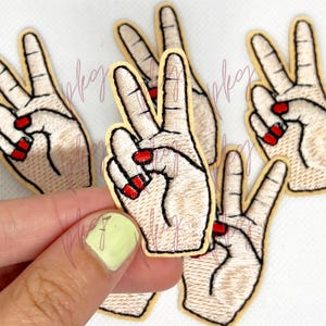 Könnte beinhalten: Gestickte Aufnäher mit einer Hand, die das Friedenszeichen zeigt. Die Hände haben rot lackierte Fingernägel und sind schwarz umrandet. Die Aufnäher haben einen beigen Hintergrund und einen gelben Rand.