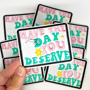 Może przedstawiać: Haftowane naszywki z napisem "HAVE THE DAY YOU DESERVE" w różowych i turkusowych literach. Naszywki mają białe tło z czarną obwódką i przedstawiają wzory stokrotek i dłoni szkieletu.