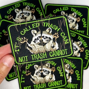 Pode incluir: Patch bordado preto com borda verde e texto que diz "CALLED TRASH CAN" e "IT'S NOT TRASH CANNOT". O patch apresenta dois guaxinins.