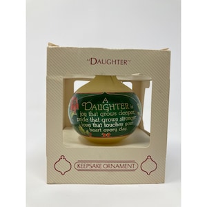 May include: A round, cream-colored glass ornament inside a beige cardboard box. The ornament features a green band with red poinsettia accents and the text: A DAUGHTER is joy that grows deeper, pride that grows stronger, love that touches your heart every day.