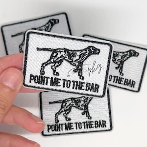 May include: Embroidered white patches with a black dog silhouette and the text "POINT ME TO THE BAR." The patches are rectangular with a black border. Several patches are visible.
