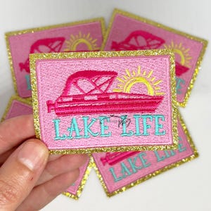 Könnte beinhalten: Rosa und gold gestickter Bügel-Patch mit einem Boot, einer Sonne und dem Text "LAKE LIFE".