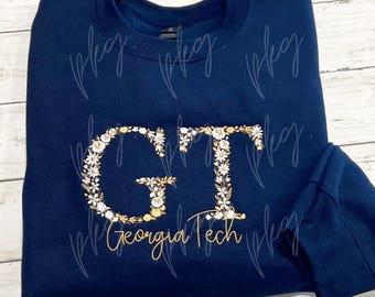 May include: Navy blue sweatshirt with floral embroidered "GT" letters and "Georgia Tech" in gold script. The floral design includes white and gold accents, creating a collegiate-themed apparel item.