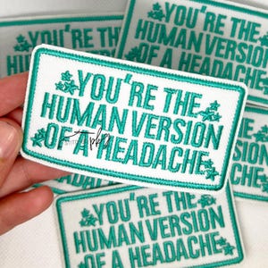 May include: White rectangular embroidered patch with a teal border and text that reads "YOU'RE THE HUMAN VERSION OF A HEADACHE". The patch has teal floral accents and is a humorous accessory.