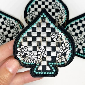 May include: Embroidered patches in a spade shape, featuring a black and white checkered pattern. The patches are outlined in black with a turquoise border and white floral accents. The patches are likely for clothing or accessories.