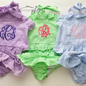 May include: Three gingham baby outfits with embroidered monograms. The outfits are in purple, green, and blue. The text "STITCHED" is visible in the top right corner of the image.