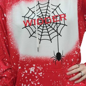 以下が含まれることがあります： 白いクモの巣のデザインと赤い「WIDDER」の文字が入った赤いパーカー。 パーカーは白い飛沫効果があり、クモの巣からぶら下がっている黒いクモが付いています。 ドローコード付きのフードが付いています。
