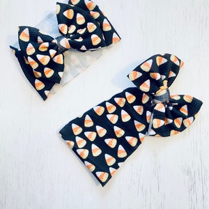 May include: Two black hair bows with a candy corn pattern. The bows are made of fabric and have a knotted center.