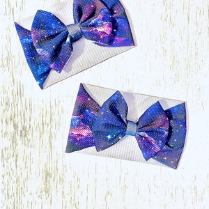 May include: Two galaxy print hair bows with a blue and purple color scheme. The bows are made of a soft, textured fabric and have a white center.