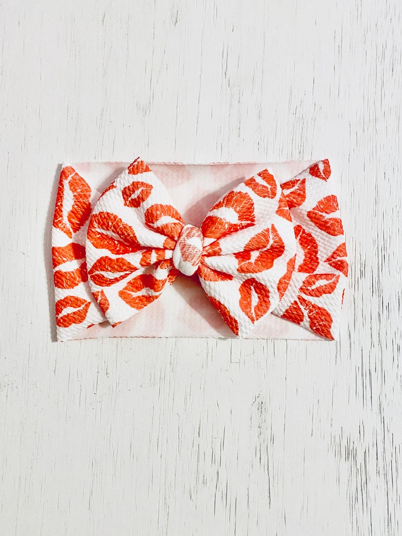 Puede incluir: Una diadema blanca con un lazo de estampado de labios rojos y blancos. El lazo est&aacute; hecho de una tela suave y texturizada.