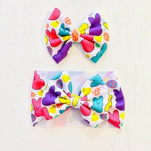 May include: Two Easter-themed hair bows with a white background and a pattern of purple, pink, yellow, and blue Peeps candy. The smaller bow is a hair clip and the larger bow is a headband.