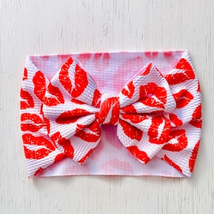 Puede incluir: Una diadema blanca con estampado de labios rojos y un gran lazo. La diadema est&aacute; atada en un lazo y tiene un efecto tie-dye rosa.