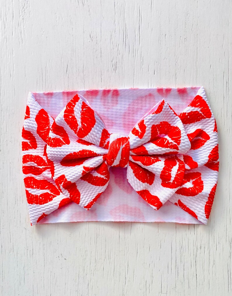 Puede incluir: Una diadema blanca con estampado de labios rojos y un gran lazo. La diadema est&aacute; atada en un lazo y tiene un efecto tie-dye rosa.