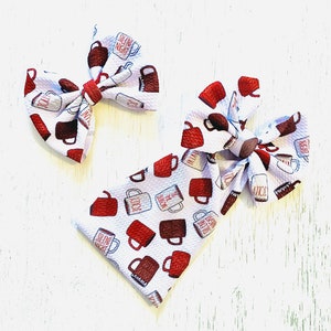May include: Two white and red hair bows with a coffee mug pattern. The mugs have different sayings on them, including "Silent Night", "Let It Snow", and "'Tis the Season".