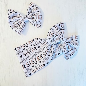 May include: Two white fabric headbands with a black and brown leopard print pattern. The headbands have the words "big sister" printed on them in a gold script font.