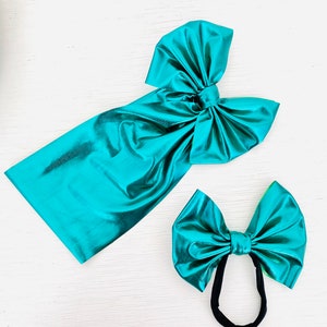 Peut inclure: Deux nœuds pour cheveux en tissu métallisé turquoise. Un nœud est grand et noué. L'autre nœud est plus petit et est attaché à un bandeau élastique noir.