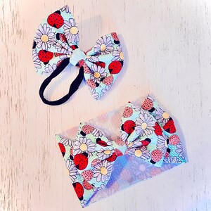 Peut inclure: Ensemble de deux accessoires pour cheveux de bébé : un bandeau et un nœud sur un élastique noir. Les deux sont décorés d'un motif répété de coccinelles rouges et de marguerites blanches sur fond bleu clair.