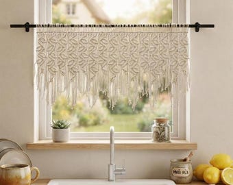 May include: A cream-colored macrame window valance featuring a repeating leaf-like knot pattern and a fringed bottom edge, suspended from a thin black horizontal rod mounted across the top of a window frame above a kitchen sink.