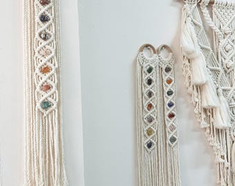 May include: Three macrame wall hangings with colorful stones woven into the design. The hangings are made with white cotton cord and have a fringe at the bottom.