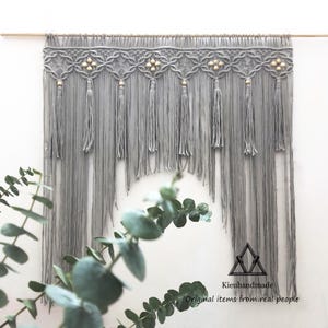 May include: A gray macrame wall hanging with a wooden rod. The design features intricate knotwork with tassels and decorative wooden beads. The bottom of the macrame has a curved shape. The text "Kieuhandmade" and "Original items from real people" is visible.