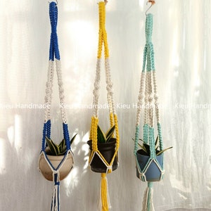 Puede incluir: Tres maceteros colgantes de macramé con cordones de diferentes colores. Los maceteros están hechos con cordones blancos y azules, blancos y amarillos, y blancos y verdes. Cada macetero tiene una pequeña planta en maceta.