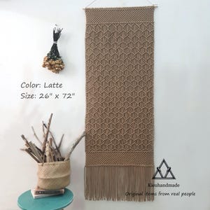 Puede incluir: Un tapiz de macramé color café con leche, de 66 cm de ancho y 183 cm de largo, con un patrón geométrico y flecos. La decoración de pared se exhibe en una pared blanca. El texto "Kieuhandmade" y "Original items from real people" es visible.