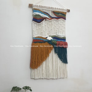 May include: A white macrame wall hanging with colorful yarn accents. The hanging features a wavy pattern with shades of blue, green, orange, and red. The hanging is finished with long white fringe.
