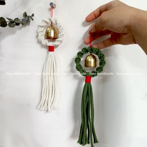 May include: Two handmade macrame bell ornaments. One features a white macrame ring, red accent, and white tassel, while the other has a green macrame ring, red accent, and green tassel. Both have a gold bell in the center and are suspended by a red string.