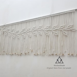 Puede incluir: Colgante de pared de macramé blanco con un diseño de hojas y cuentas de madera. El colgante está hecho con cordón de algodón y tiene una franja en la parte inferior.