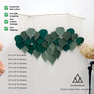 May include: A wall hanging made of green felt leaves, arranged in a cascading pattern. The leaves are attached to a white string, which is hung from a white rod. The wall hanging is available in various sizes, with the largest measuring 120 cm tall by 110 cm wide. The wall hanging is handmade and features the text "Kieuhandmade Original items from real people".