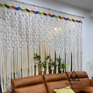 May include: Handmade macrame curtain in off-white with a colorful top border. The curtain features a leaf pattern and long, flowing strands. The design is displayed in front of a window, with a brown leather sofa in the foreground.