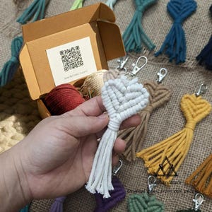 May include: A white macrame heart keychain with a silver clasp is held in front of a box containing red and beige macrame cord. Other colorful macrame keychains are visible in the background. The box has a QR code and text.