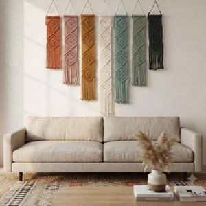 May include: Seven vertical macrame wall hangings in a row, featuring colours of burnt orange, dusty rose, mustard yellow, cream, teal, sage green, and black. Each piece displays intricate knotted geometric patterns and long fringe at the bottom.