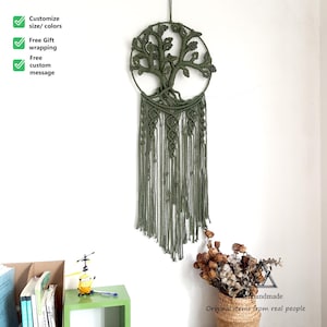 Op de afbeelding: Een handgemaakte olijfgroene macramé wandhanger met een levensboomontwerp. Het cirkelvormige frame bevat de boom, met ingewikkelde knopen en lange franjes. De wanddecoratie wordt tegen een witte muur getoond.