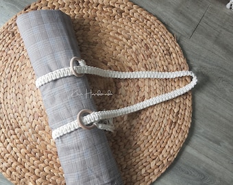 May include: A rolled up plaid fabric yoga mat with a white cotton carrying strap and wooden ring handles. The mat is lying on a woven straw mat.