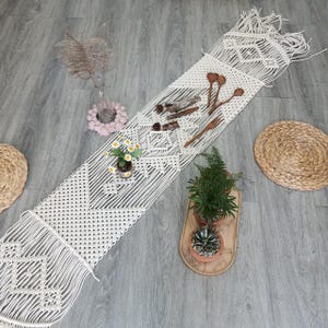May include: A white macrame table runner with a geometric pattern. The runner is laid out on a grey wood floor with potted plants and wooden spoons on top.