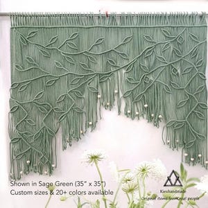 Pode incluir: Uma tapeçaria de macramé em verde sálvia com um padrão de videiras e folhas, decorada com pequenas contas de madeira nas franjas. O texto na imagem diz: Shown in Sage Green (88,9 cm x 88,9 cm), Custom sizes & 20+ colors available, Kieuhandmade.