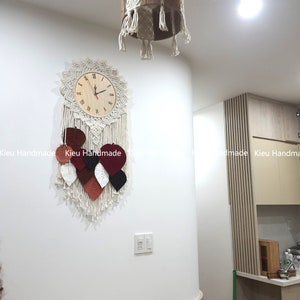 May include: A white macrame wall hanging with a wooden clock in the centre. The clock has Roman numerals and black hands. There are eight felt hearts hanging from the bottom of the macrame, in shades of red, brown, and white.