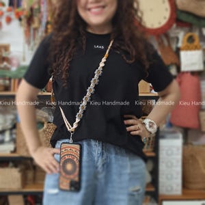 Puede incluir: Una persona lleva una camiseta negra con la palabra "LAAS" impresa en ella y pantalones vaqueros azules. Lleva una correa de teléfono con un diseño floral y una funda de teléfono con un patrón colorido.