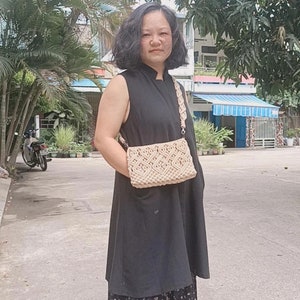 May include: A woman wearing a black sleeveless dress with a floral print underskirt and a beige crocheted crossbody bag. She is standing on a paved surface.