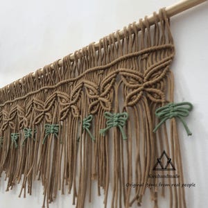 May include: A handmade macrame wall hanging in shades of brown and green. The design features repeating floral patterns and knotted details, with a wooden dowel at the top for hanging. The bottom has long fringe. The text "Kieuhandmade Original items from real people" is visible.