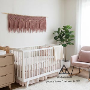May include: A nursery scene featuring a white crib with pink bedding and a matching skirt. Above the crib hangs a dusty rose macrame wall hanging with wooden accents. A pink rocking chair and a dresser complete the room.
