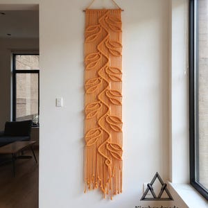 May include: Orange macrame wall hanging featuring a leaf design. The artwork is rectangular and hangs vertically, with a wooden dowel at the top. The leaves are intricately knotted, and the bottom has fringe. The text "Kieuhandmade" and "Original items from real people" are at the bottom.