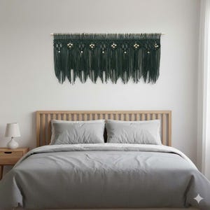 May include: Dark green macrame wall hanging with a fringe design and wooden beads. The macrame is mounted on a wooden dowel. The wall hanging is above a bed with gray bedding.