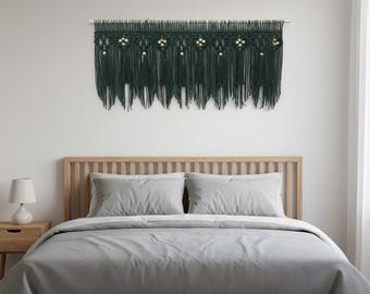 May include: Dark green macrame wall hanging with a fringe design and wooden beads. The macrame is mounted on a wooden dowel. The wall hanging is above a bed with gray bedding.