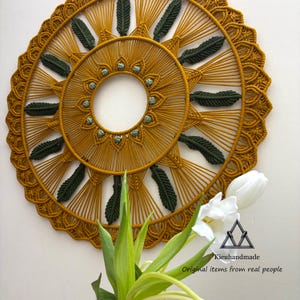 May include: A circular macrame wall hanging featuring a mustard yellow cord design with dark green leaf-shaped accents and small light green beads arranged in a ring around the center. The piece includes a logo with the text Kieuhandmade.