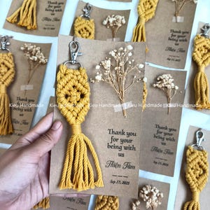 Puede incluir: Un llavero de macramé color amarillo mostaza con patrón de diamante y borla sujeto a una tarjeta marrón. La tarjeta presenta una ramita de flor blanca seca y el texto impreso: Thank you for your being with us Thien Tam Sep 17, 2023.