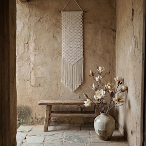 May include: A cream-colored macrame wall hanging with a geometric pattern and fringe hangs on a textured wall. A rustic wooden bench and a vase of faux flowers complete the scene, creating a bohemian aesthetic.