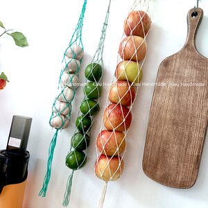 May include: Three hanging produce bags with different fruits and vegetables. The bags are made of white and green string. The bags are hanging from a white wall. The bags are filled with red apples, green limes, and white garlic.