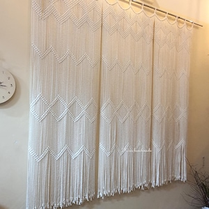 Puede incluir: Cortinas de macramé color crema con patrones geométricos de diamantes y zig-zag. Las cortinas cuelgan de una barra plateada, añadiendo un toque bohemio a la habitación. El diseño presenta detalles de cuerda anudada y un dobladillo con flecos.